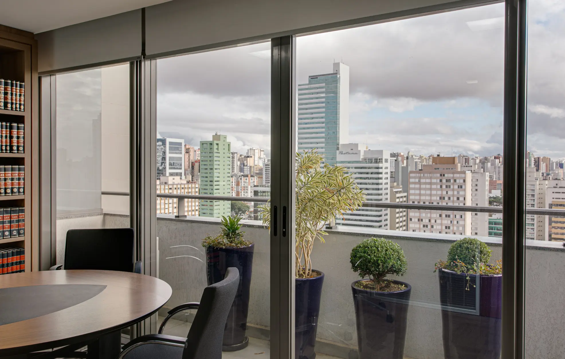 Sala de atendimento com vista para a cidade de Curitiba, sacada com plantas, mesa redonda de madeira e estante com livros de Direito.