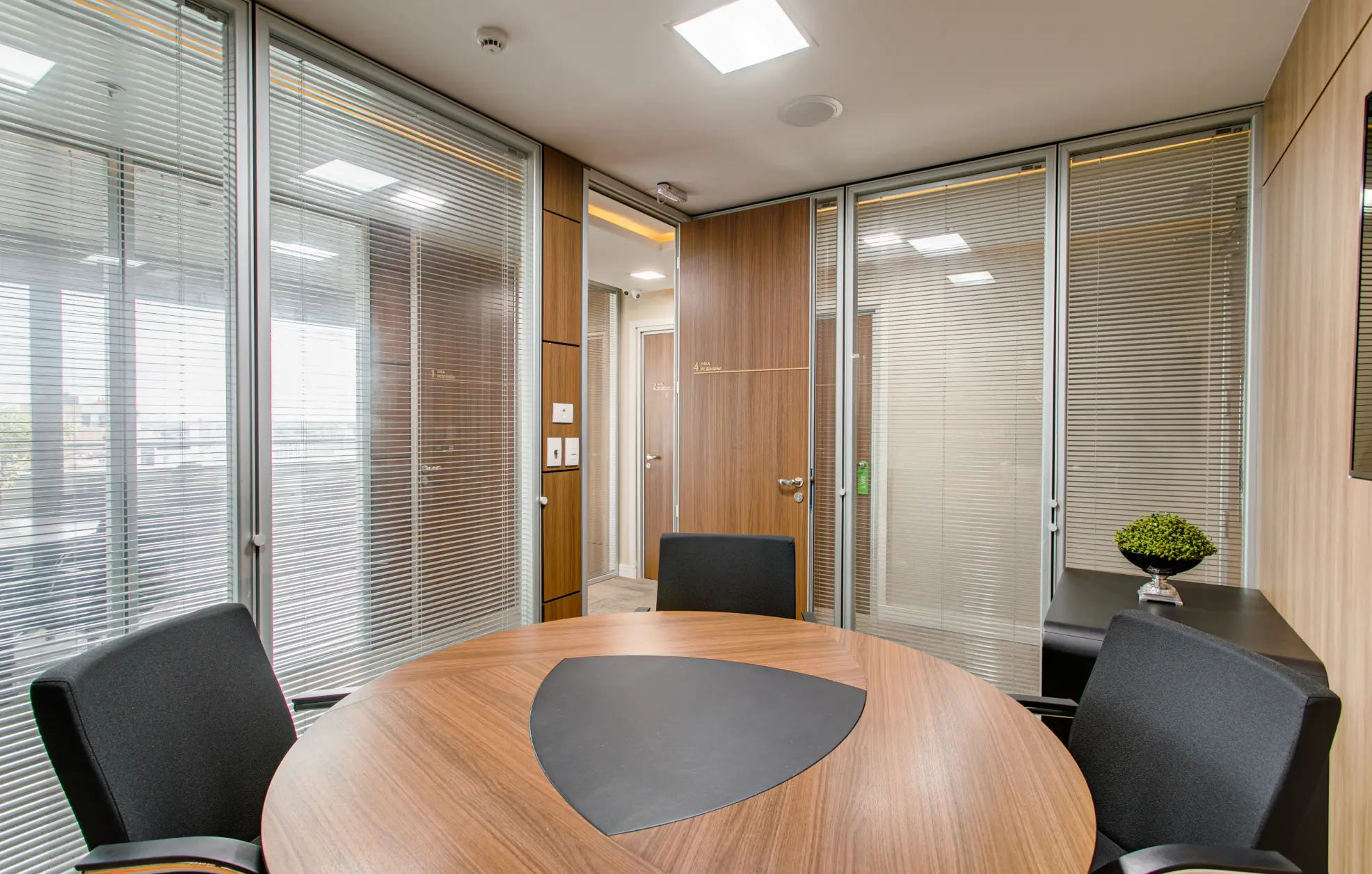 Sala de atendimento interna com paredes de vidro e persianas, mesa redonda de madeira e balcão preto com planta.