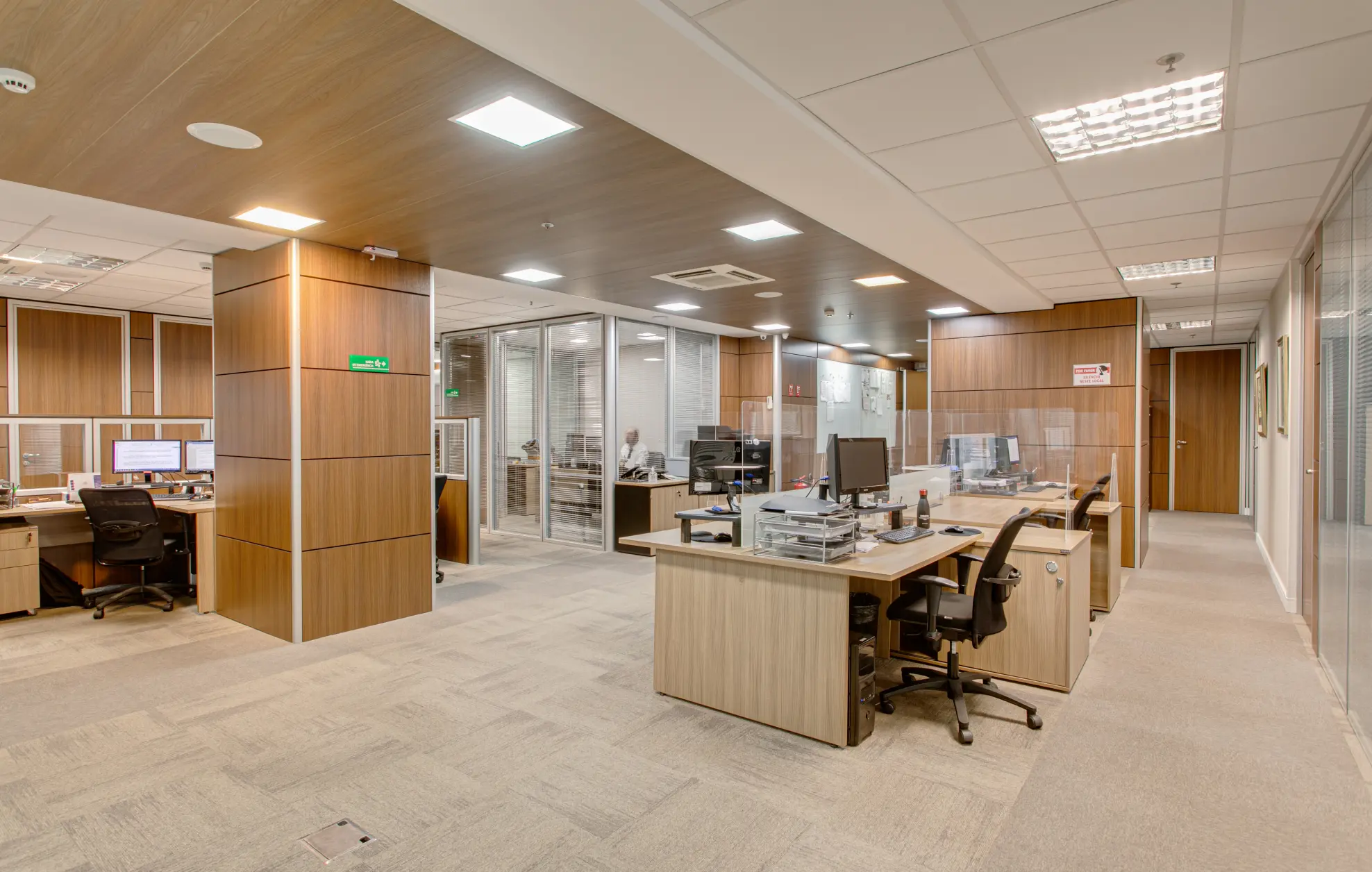 Sala ampla com painéis de madeira, piso em carpet cinza, mesas com computadores e outras salas em volta.